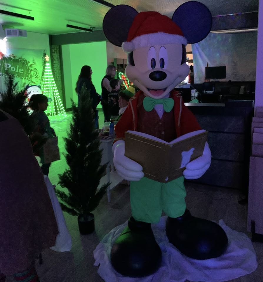 A life-size Mickey Mouse caroling decoration in a Santa hat, holding a songbook.