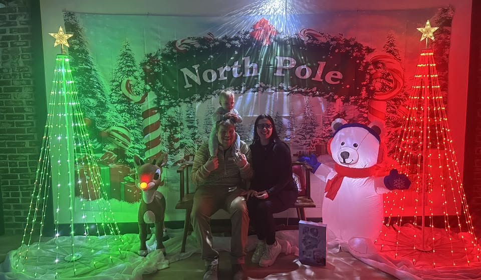A family posing for a photo in front of the North Pole backdrop, with lit Christmas trees and a Rudolph decoration.
